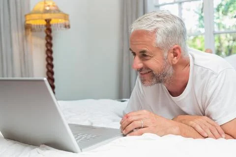 Senior man lying on bed propped on elbows, looking at silver laptop and wearing Stock Photos