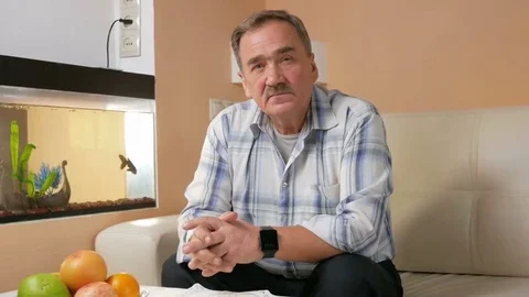 Senior man mustache thoughtfully looking at the camera. He sits on the couch at Stock Footage 69212684