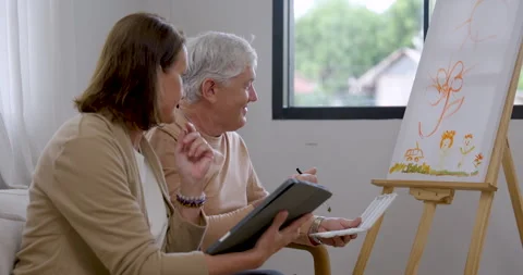 Senior man paints flower artwork, pauses to view tablet with senior woman Stock Footage 340169199