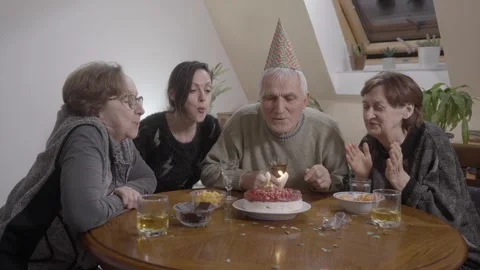 Senior man an party hat sitting at dinner table blowing candles on Birthday cake Stock Footage 331315824