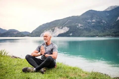 A senior man pensioner sitting by lake in nature, doing yoga exercise. Stock Photos