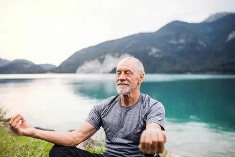 A senior man pensioner sitting by lake in nature, doing yoga exercise. Stock Photos
