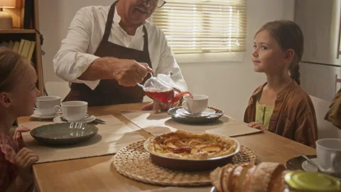 Senior Man Pouring Hot Tea for Granddaughters at Kitchen Table Stock Footage 344551409