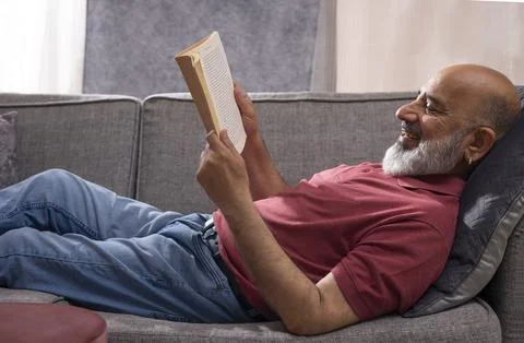 Senior man reading a book while lying on sofa at home Stock Photos