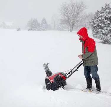 Senior man in red coat using a snow blower during a blizzard on home drive... Stock Photos