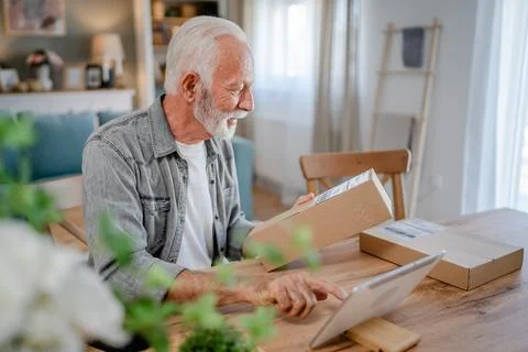 Senior man sit at home hold post package delivery use digital tablet Stock Photos