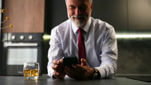 Senior man sit in kitchen drinking whiskey using smartphone, web surfing Stock Footage 234650370