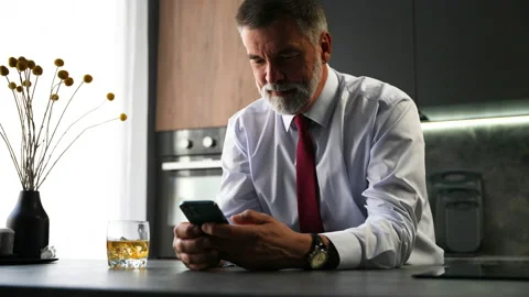 Senior man sit in kitchen drinking whiskey using smartphone, web surfing Video stock 234650590