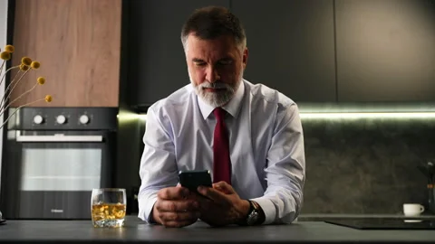 Senior man sit in kitchen drinking whiskey using smartphone, web surfing Video stock 234650613