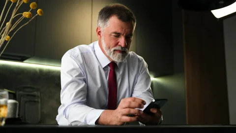 Senior man sit in kitchen drinking whiskey using smartphone, web surfing Video stock 234650738