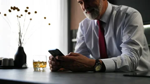 Senior man sit in kitchen drinking whiskey using smartphone, web surfing Video stock 234650757