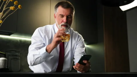 Senior man sit in kitchen drinking whiskey using smartphone, web surfing Video stock 234650972