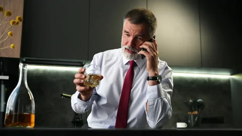 Senior man sit in kitchen drinking whiskey talking on smartphone Stock Footage 234650999