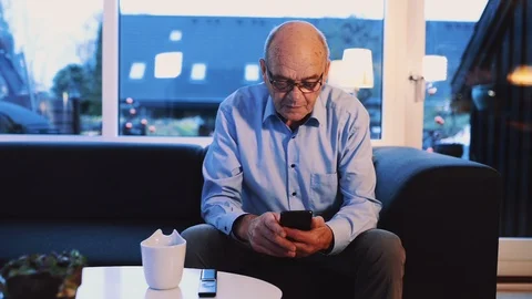 Senior man sits in sofa looking at the screen of his smart phone Stock Footage 99013456