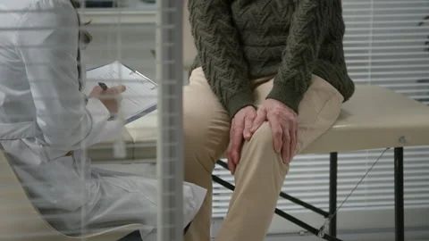 Senior Man Sitting on Exam Table Complaining about Knee Pain at Doctors Office Stock Footage 333203763