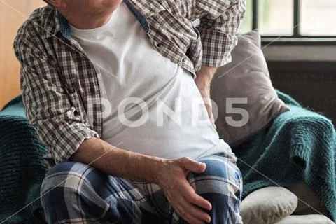 Senior man sitting indoor on the armchair feeling sudden ache in his back Stock Photos
