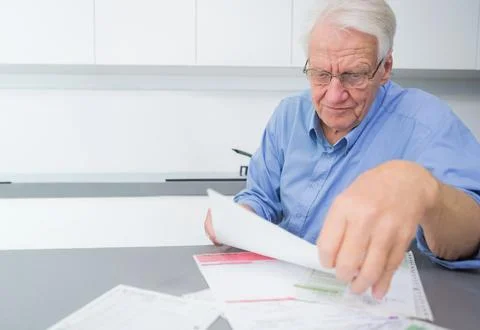 Senior man sitting at kitchen table sorting paperwork, wearing glasses and blue Stock Photos