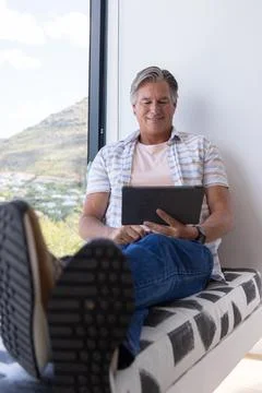 Senior man sitting on patterned bench by large vertical window, holding tablet Stock Photos