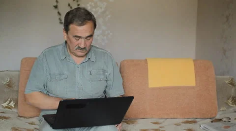 Senior man sitting on sofa with laptop at home. He carefully works at the Stock Footage 65374493