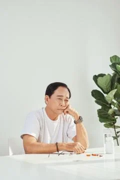 Senior man sitting at table, having bad headache, grimacing, taking medicine. Stock Photos