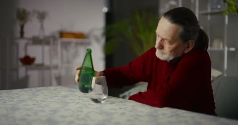 Senior man sitting at a table holding a green bottle in a quiet home interior Video stock 336289320