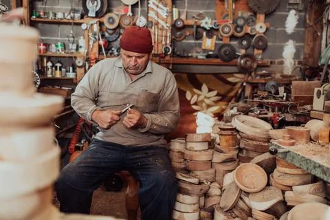 A senior man sitting in the workshop and processing wooden utensils in the old Stock Photos