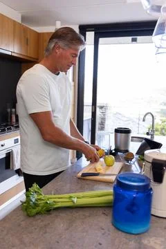 Senior man standing at kitchen counter slicing banana on wooden cutting board Stock Photos