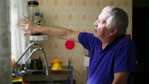 Senior man standing by kitchen window peeking out through curtain contempla.. Stock Photos