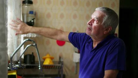 Senior man standing by kitchen window peeking out through curtain contempla.. Stock Photos