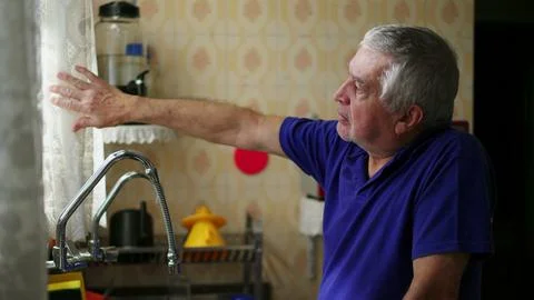 Senior man standing by kitchen window peeking out through curtain contempla.. Stock Photos