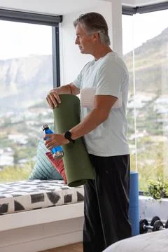 Senior man standing by large windows holding green yoga mat, bottle and towel Foto stock