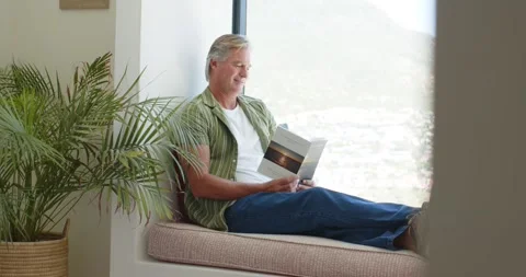 Senior man starting upright holding open book, sitting on window seat, reading Stock Footage 340973695