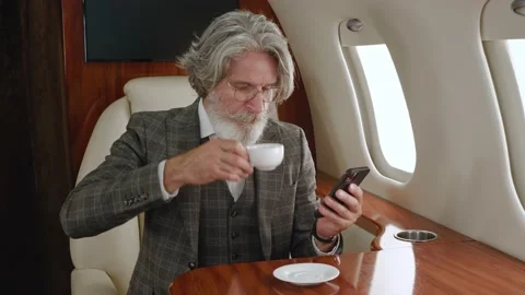 Senior man in suit using smartphone and drinking tea while flying in private Stock Footage 198084153