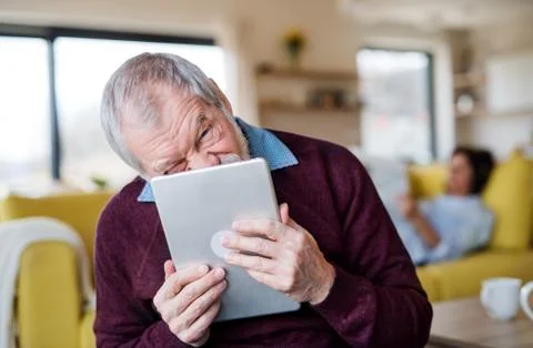 Senior man with tablet indoors at home, making funny faces. Stock Photos