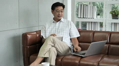Senior man thinking, sitting on couch in living room, using laptop for online 스톡 동영상 138306247