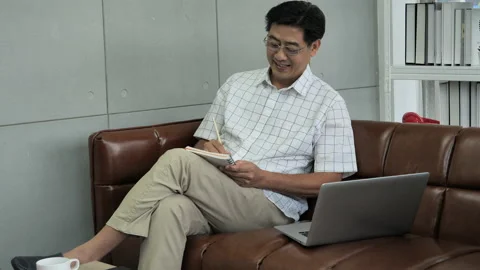 Senior man thinking, sitting on couch in living room, using laptop for online 스톡 동영상 138306599