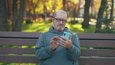 Senior man uses a smartphone while sitting on a park bench. Pensioner surfing Stock Footage 264323821