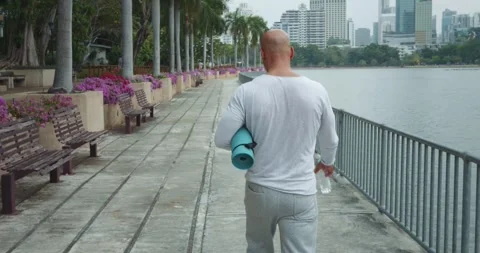Senior man walking through public park with yoga mat hand seeking perfect place Stock Footage 274429151