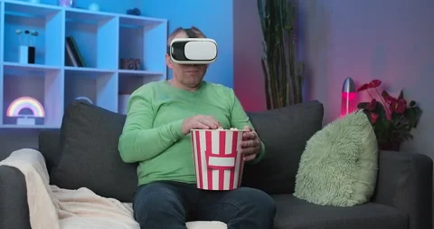 Senior man wearing virtual reality glasses eating popcorn while sitting on sofa Stock Footage 194476199