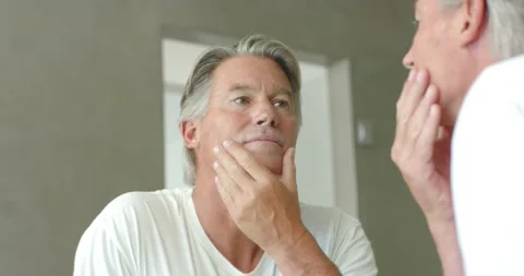 Senior man wearing white tee checking skin and jawline in bathroom mirror upon Stock Footage 337160406