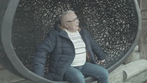 Senior man in winter jacket sits in hanging cocoon chair Stock Footage 148388799