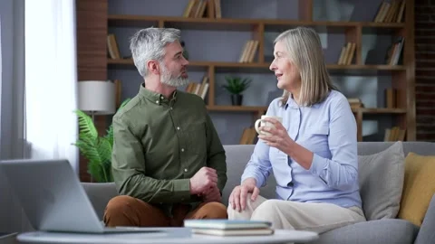 Senior married couple talking over coffee while sitting on a sofa in the living  Stock Footage 339775627