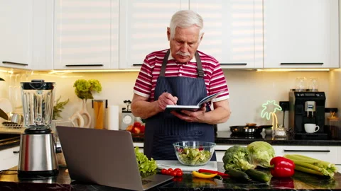Senior old man making vegetable salad using laptop recipe and writing notebook Stock Footage 347034782