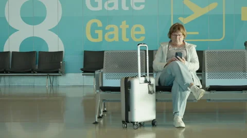 Senior passenger sits with suitcase and checks smartphone at airport gate. Stock Footage 343984121