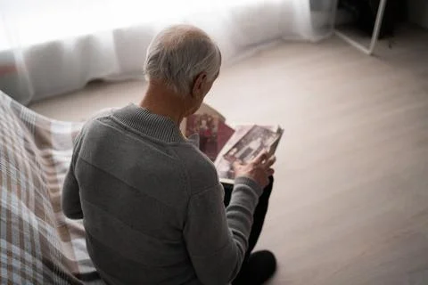 Senior sad man looking old photo missing his wife sitting on sofa at home 库存照片