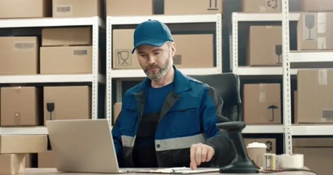 Senior warehouse worker in uniform sitting at desk folds cardboard boxes Stock Footage 150383338