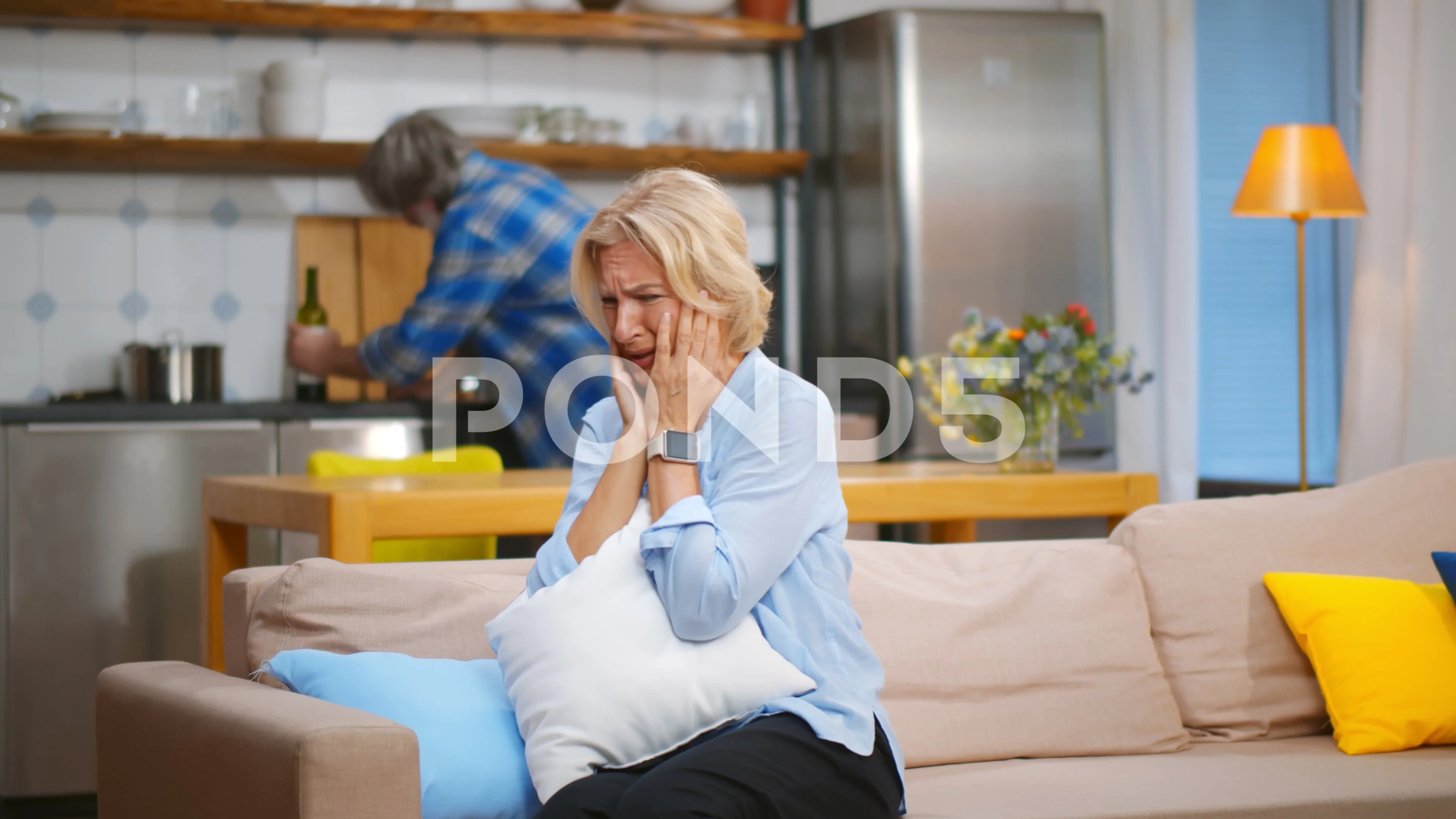Senior wife sitting on couch and crying with husband drinking on background