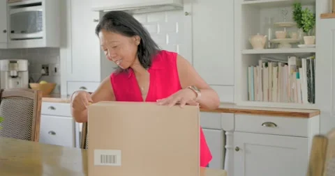 Senior woman is approaching dining table and is cutting tape on cardboard box to Stock Footage 345037496