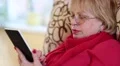 Senior woman with eyeglasses sits on a sofa and reads electronic book HD Footage