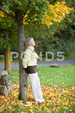 Senior woman leaning on tree trunk in autumn at park Stock Photos